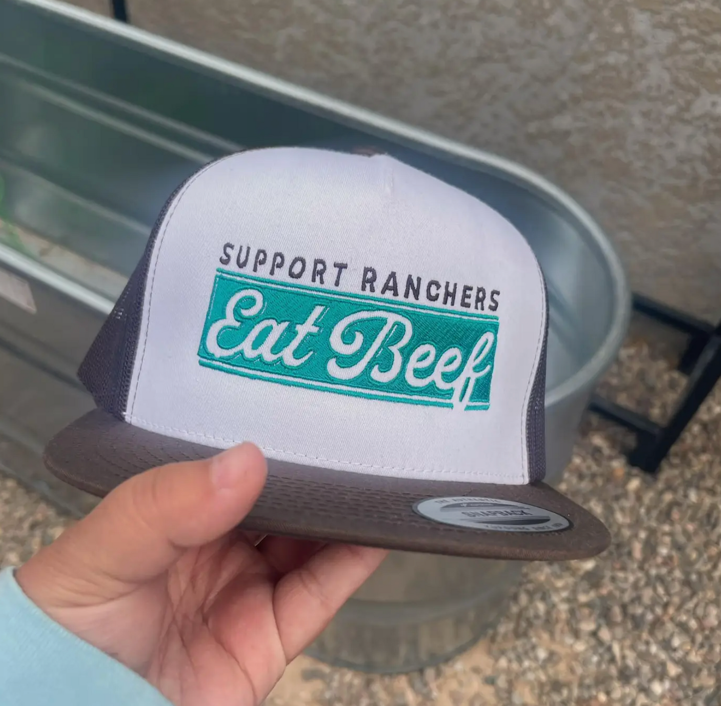 Trucker hat with 'Support Ranchers Eat Beef' text held by a hand.