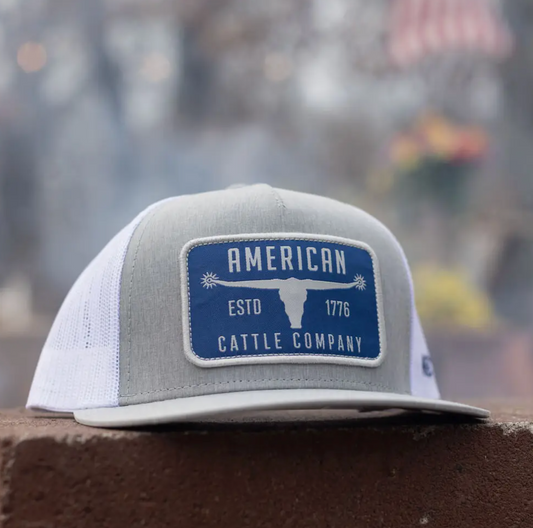 Gray and white cap with a blue patch featuring 'American Cattle Company' on a blurred background.