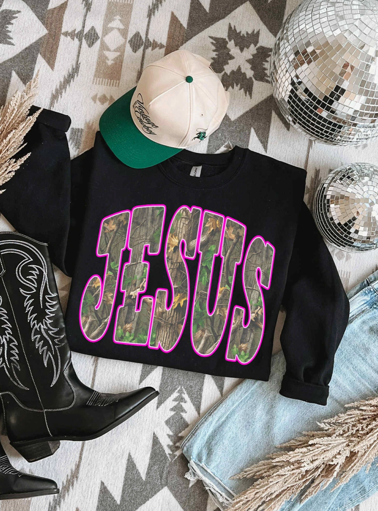 Black sweatshirt with 'Jesus' printed on it, surrounded by cowboy boots, a cap, and disco balls on a patterned rug.
