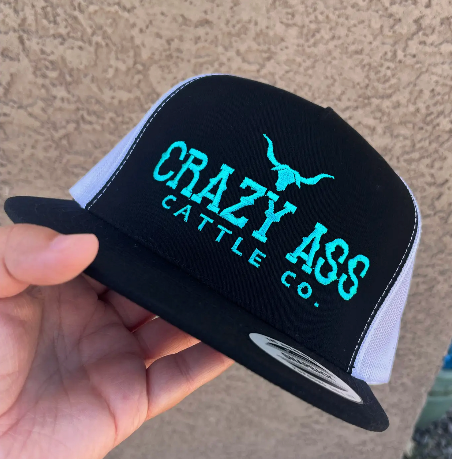 Black and white cap with 'Crazy Ass Cattle Co.' text held by a hand against a beige wall.
